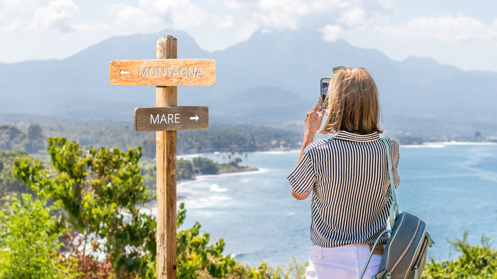 Zdjęcie przedstawia młodą dziewczynę z włosami do ramion. Dziewczyna ubrana jest w koszulę w paski i spodnie. Na plecach nosi mały plecak. Dziewczyna stoi tyłem do kadru, niewidoczna jest jej twarz. Dziewczyna wykonuje telefonem zdjęciem krajobrazu widzianego z góry. Na zdjęciu widoczne jest morze, a w oddali szczyty gór. Obok dziewczyny, po lewej stronie znajduje się drewniany drogowskaz z dwoma tabliczkami. Na górnej tabliczce jest napis Montagna oraz strzałka w lewo, na dolnej tabliczce jest napis Mare oraz strzałka w prawo. Na zdjęciu obok drogowskazu widoczna jest również górska roślinność.