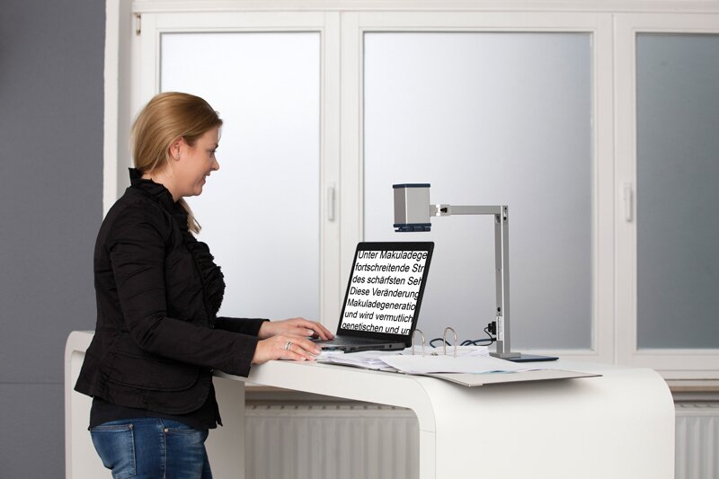 Ilustracja przedstawia uśmiechniętą kobietę stojącą przy biurku, na którym leży otwarty laptop. Kobieta wpatrzona jest w ekran, trzyma dłonie na klawiaturze. Na wyświetlaczu widać tekst o foncie dużo większym od standardowego.