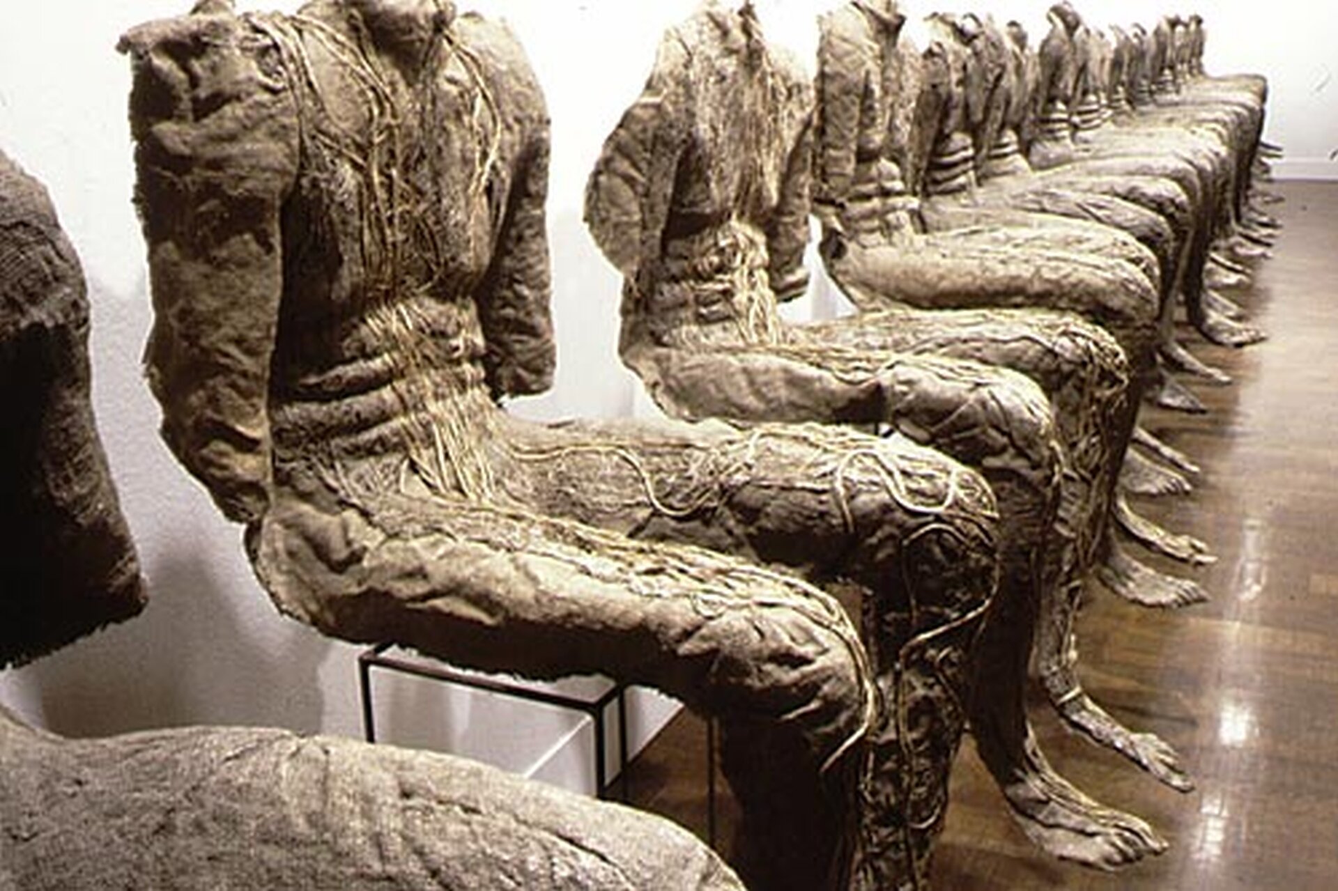 Ilustracja przedstawia instalację artystyczną składającą się z siedzących postaci, które nie mają głowy. Ręce postaci kończą się na wysokości łokcia. Stopy nie dotykają podłogi, zwisają nad nią.