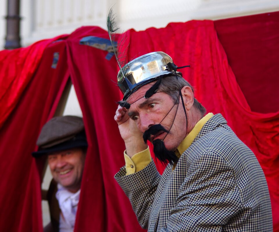 Fotografia przedstawia mężczyznę. Postać ukazuje lewy profil. Mężczyzna ma na głowie rondel z wetkniętym weń pawi piórem. Rondel ma tasiemkę założoną na brodę mężczyzny. Do tasiemki umocowano sztuczne brwi, wąsy i hiszpańską brodę. Postać marszczy czoło i pochyla głowę w lewą stronę. Jest ubrany w koszulę i kraciastą marynarkę. Za jego plecami wisi kurtyna. Wygląda zza niej uśmiechnięty mężczyzna.