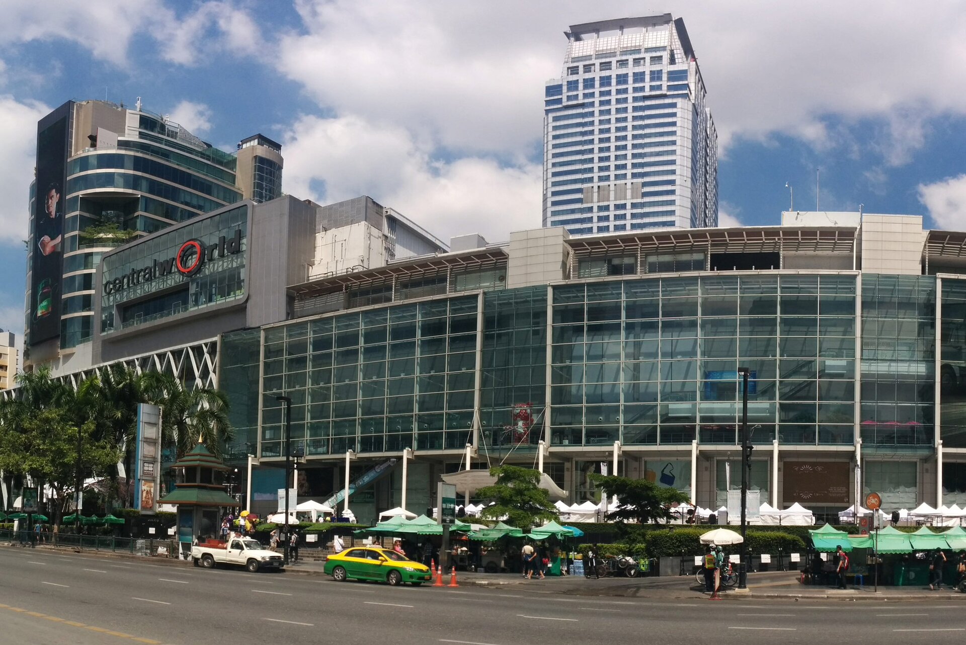 Zdjęcie przedstawia nowoczesny, przeszklony budynek składający się z niskiej części biegnącej po lekkim łuku oraz wyżej, wielopiętrowej części znajdującej się po lewej stronie. Na tej partii budynku, na elewacji, widoczny jest napis: central world. Przed budynkiem widoczny jest szeroki chodnik oraz ulica. Na chodniku widoczne są rozstawione namioty i markizy w kolorze zielonym i białym, pod którymi znajdują się stoiska handlowe. W tle widoczne są dwa wielopiętrowe budynki górujące nad budynkiem centrum handlowego.