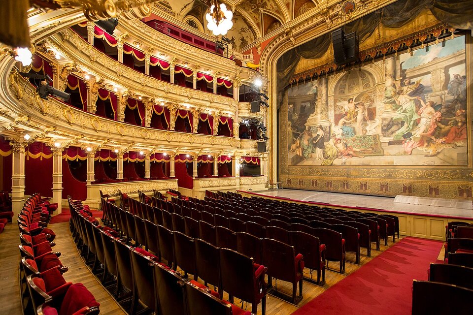 Zdjęcie przedstawia salę teatralną. Po lewej stronie pomieszczenia znajdują się widownia oraz loże, a po prawej scena z kurtyną przedstawiającą antyczną scenę rodzajową. W wystroju sali dominują kolory złoty i czerwony.  