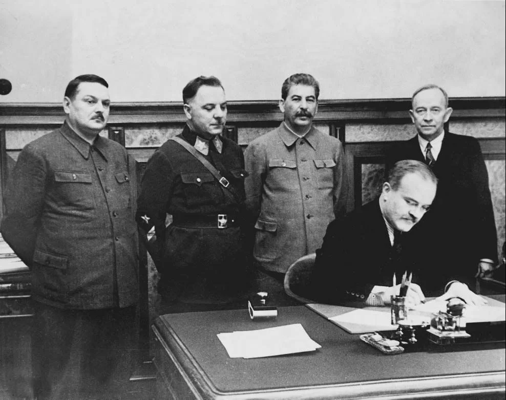 Kliknij, aby powiększyć Zdjęcie przedstawia Mołotowa w garniturze siedzącego przy biurku i podpisującego dokumenty. Za jego plecami stoi Stalin w wojskowym uniformie w towarzystwie trzech mężczyzn - po lewej stronie dwóch ubranych w wojskowe uniformy, po prawej Kuusinen w garniturze.
