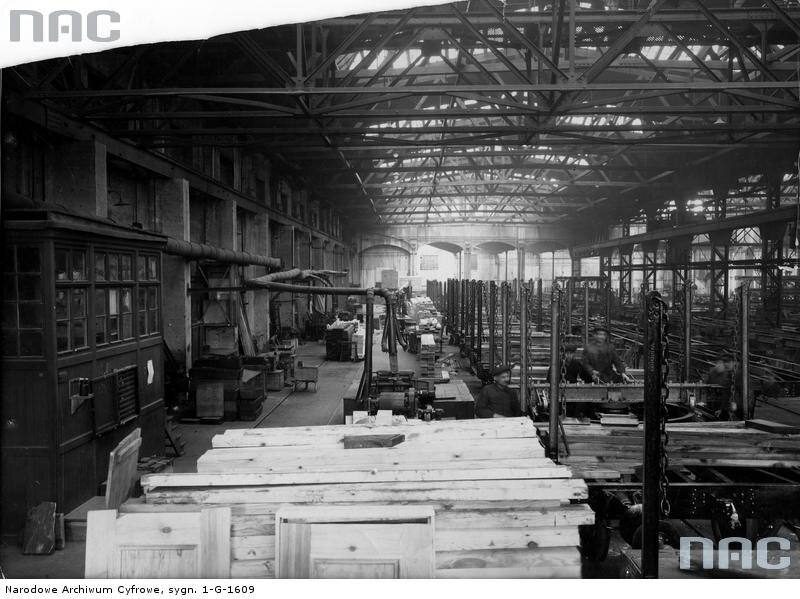 Czarno‑białe zdjęcie przedstawia wnętrze hali fabryki. Przy maszynach i stanowiskach pracują liczni robotnicy. Przy ścianach hali stoją szafki z licznymi narzędziami. W dachu znajdują się szyby, przez które do hali wpada światło. 
