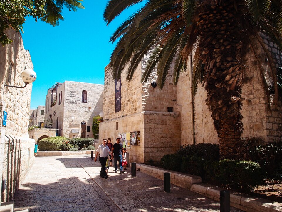 Zdjęcie przedstawia dzielnicę żydowską w Jerozolimie. W centrum jest szeroki chodnik, z obu stron otaczają go wysokie kamienne mury budynków. Po chodniku spacerują ludzie. Na pierwszym planie palma.