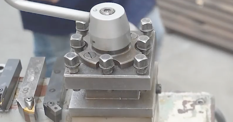 Fotografia przedstawia imak nożowy. Jest to przyrząd służący do zamocowywania narzędzi na obrabiarkach w postaci prostokątnej płytki z trzema otworami wzdłuż każdej krawędzi ze śrubami oraz trzpieniem znajdującym się w środku, na przecięciu dwóch przekątnych, do którego przymocowana jest nakładka z odchodzącym w bok metalowym ramieniem. 