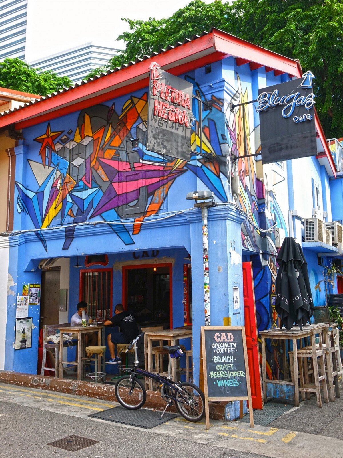 Zdjęcie przedstawia graffiti namalowane na fasadzie niebieskiego budynku z czerwonym dachem. Graffiti ukazuje kolorowe wzory geometryczne. Przed budynkiem stoi rower. Budynek ze zdjęcia to najprawdopodobniej restauracja. Przed budynkiem stoją stoliki oraz krzesła, na których siedzą dwie postacie.