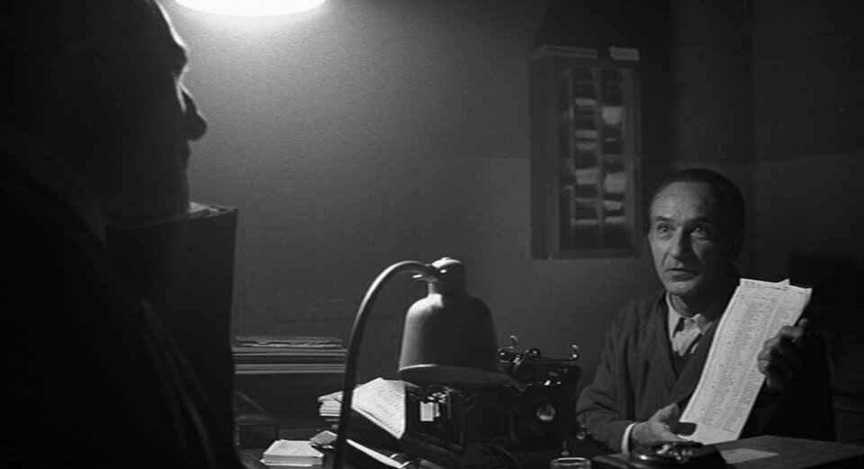 Zdjęcie przedstawia wnętrze pokoju. Jest w nim Schindler i naprzeciw niego mężczyzna w garniturze i okularach. Trzyma w dłoniach kartki papieru z listą. Pokazuje je Schindlerowi.