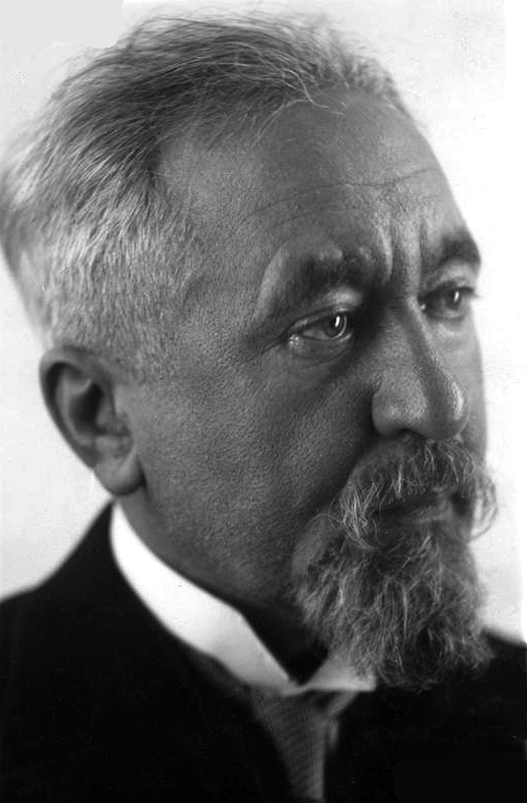 Zdjęcie przedstawia mężczyznę z zarostem i siwymi włosami. Ma poważny wyraz twarzy. Spod jego marynarki wystaje kołnierzyk koszuli. 