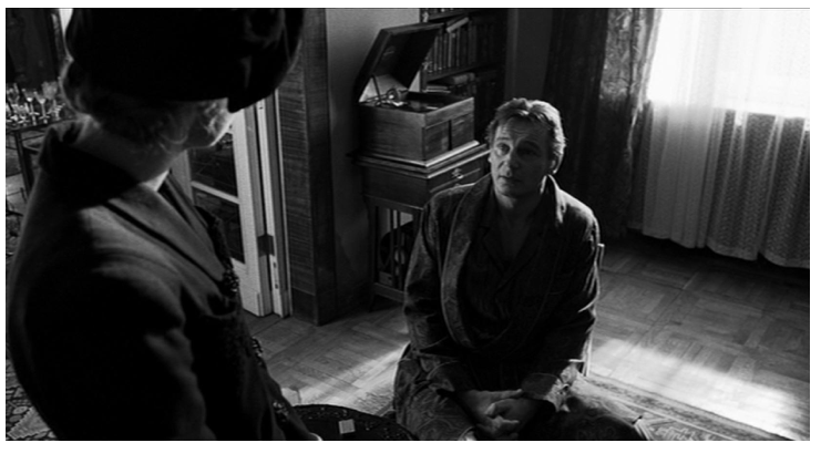 Zdjęcie przedstawia wnętrze pokoju. Na podłodze klęczy Schindler. Jest w szlafroku. Przed nim stoi kobieta w płaszczu i kapeluszu.