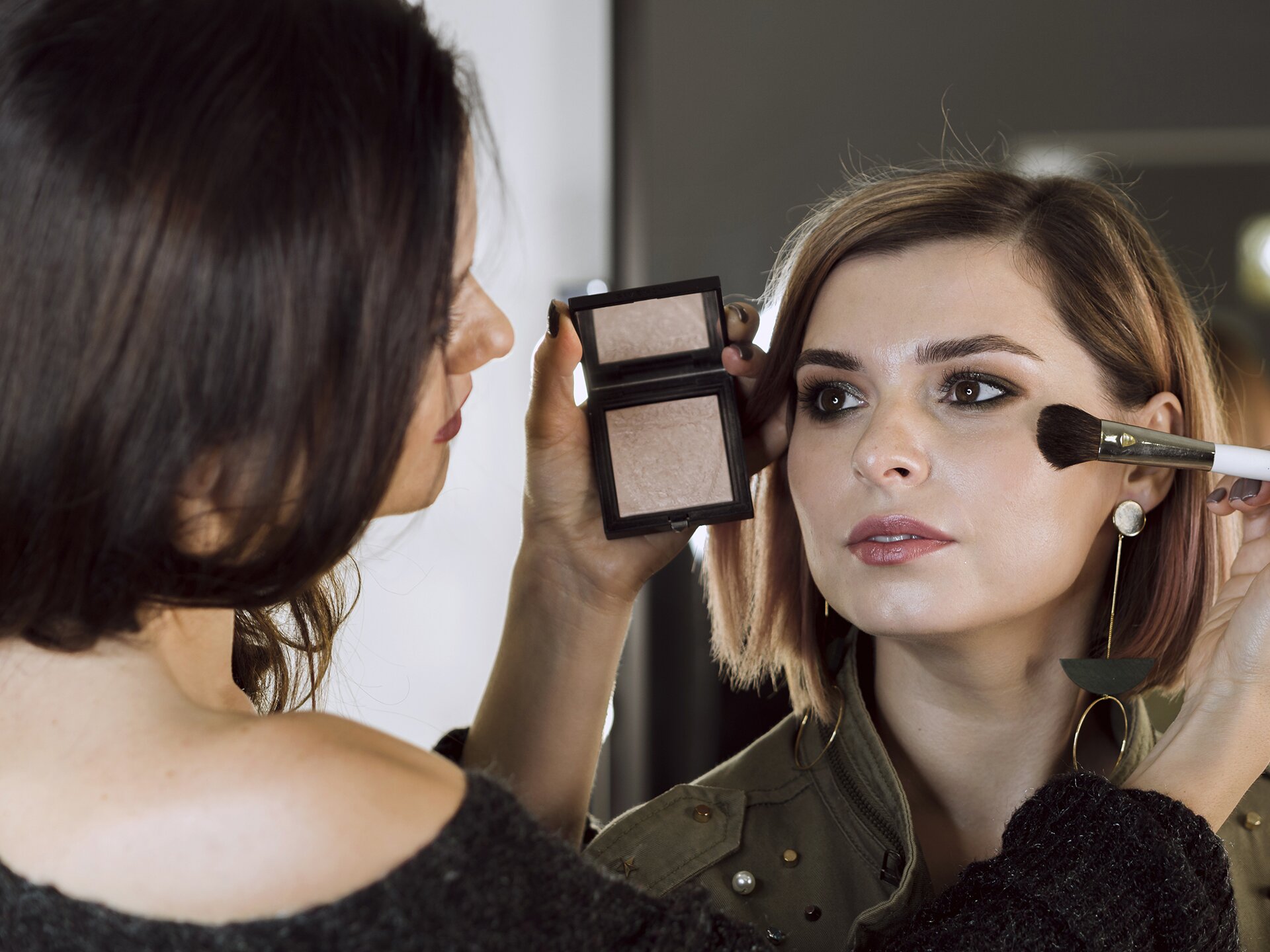 Zdjęcie przedstawia makijażystkę wykonującą makijaż kobiecie. Używa pudru i pędzelka do nakładania kosmetyku na twarz.