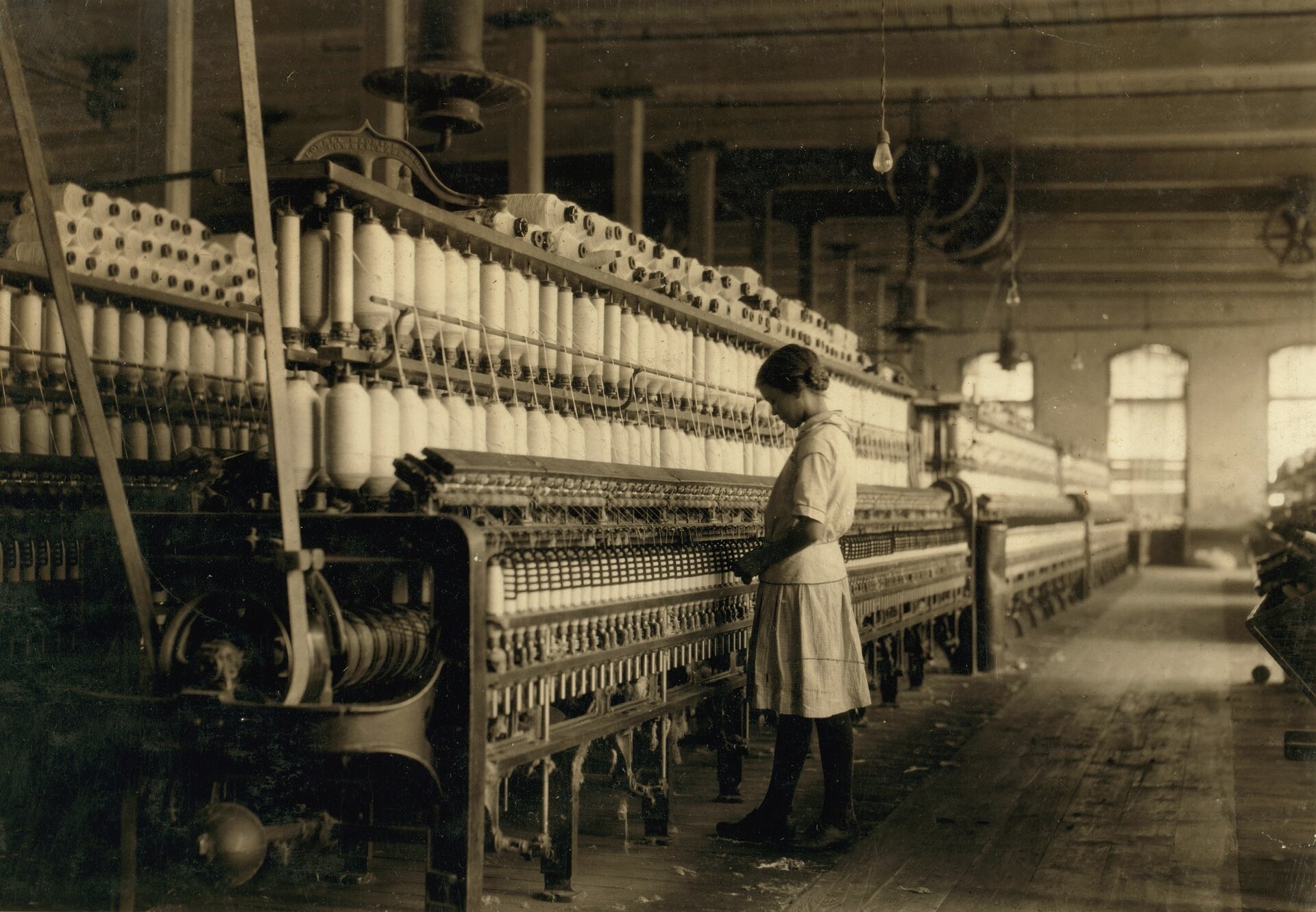 Zdjęcie przedstawia dziewczynkę, która stoi przed maszyną włókienniczą w hali. Na maszynie umieszczono wiele szpul z nicią. 