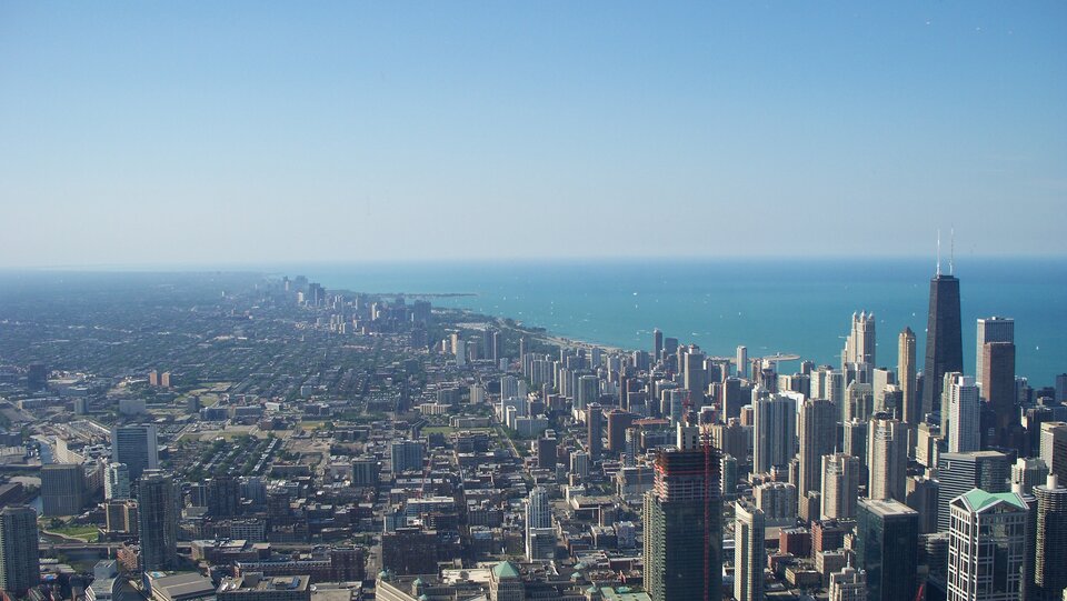 Na fotografii widoczne jest Chicago z lotu ptaka. Miasto posiada gęstą zabudowę składającą się z drapaczy chmur i niższych budynków. Po prawej stronie zdjęcia znajduje się Jezioro Michigan.