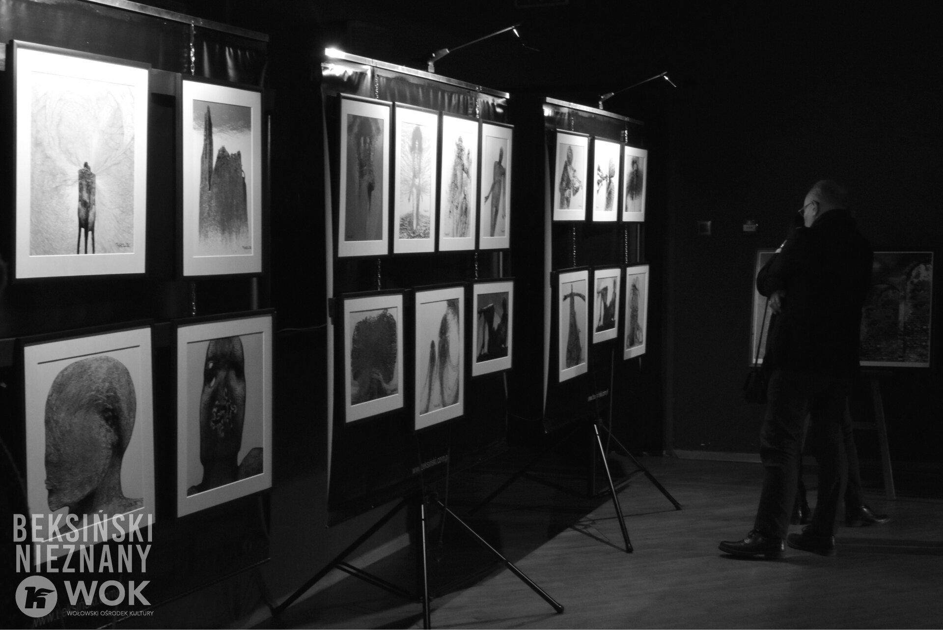 Czarno‑białe zdjęcie przedstawia fragment wystawy. Na wysokich sztalugach rozmieszczono prace artysty – fotografie, grafiki oraz fotomontaże. Na pierwszej sztaludze zaprezentowano cztery dzieła, na drugiej - siedem, a na trzeciej – sześć. Przy trzeciej sztaludze stoi mężczyzna podziwiający prace. W lewym dolnym rogu umieszczony został znak wodny: BEKSIŃSKI NIEZNANY WOK – WOŁOWSKI OŚRODEK KULTURY.