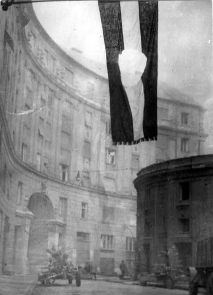 Zdjęcie przedstawia miejską ulicę. Nad ulicą powiewa węgierska flaga. Ma ze środka wycięty okrągły fragment.