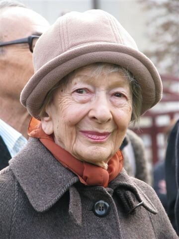 Zdjęcie przedstawia portret starszej kobiety. Jej twarz jest szczupła, widać na niej liczne zmarszczki. Kobieta uśmiecha się. Ubrana jest w płaszcz, pod szyją ma zawiązaną apaszkę, a na głowie ma założony niewielki kapelusz.