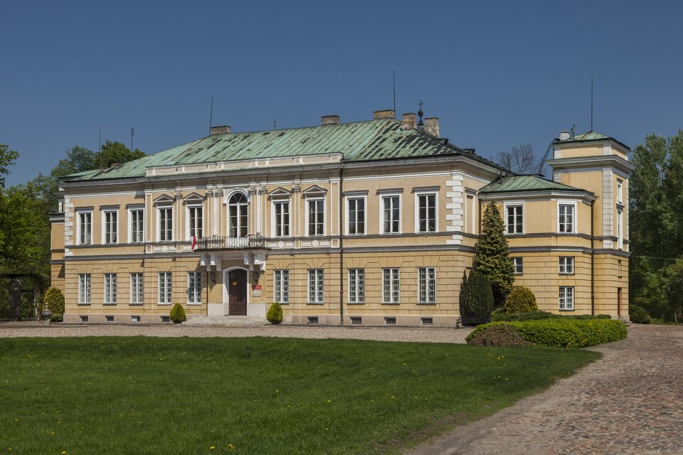 Ilustracja przedstawia Pałac Prymasowski w Skierniewicach. Jest to budowla dwukondygnacyjna z wysuniętą częścią środkową. Posiada czterospadowy dach z dachówkami. Parter jest wydzielony gzymsem. Okna są obramione, nad nimi znajduje się belkowanie. Po obu stronach wejścia są po dwie kolumny. Ryzalit wejściowy jest zwieńczony balkonem, nad którym jest portal zamknięty półkoliście, flankowany zdwojonymi pilastrami dźwigającymi przełamujące się belkowanie. Obok pałacu trawa z różnymi gatunkami drzew.