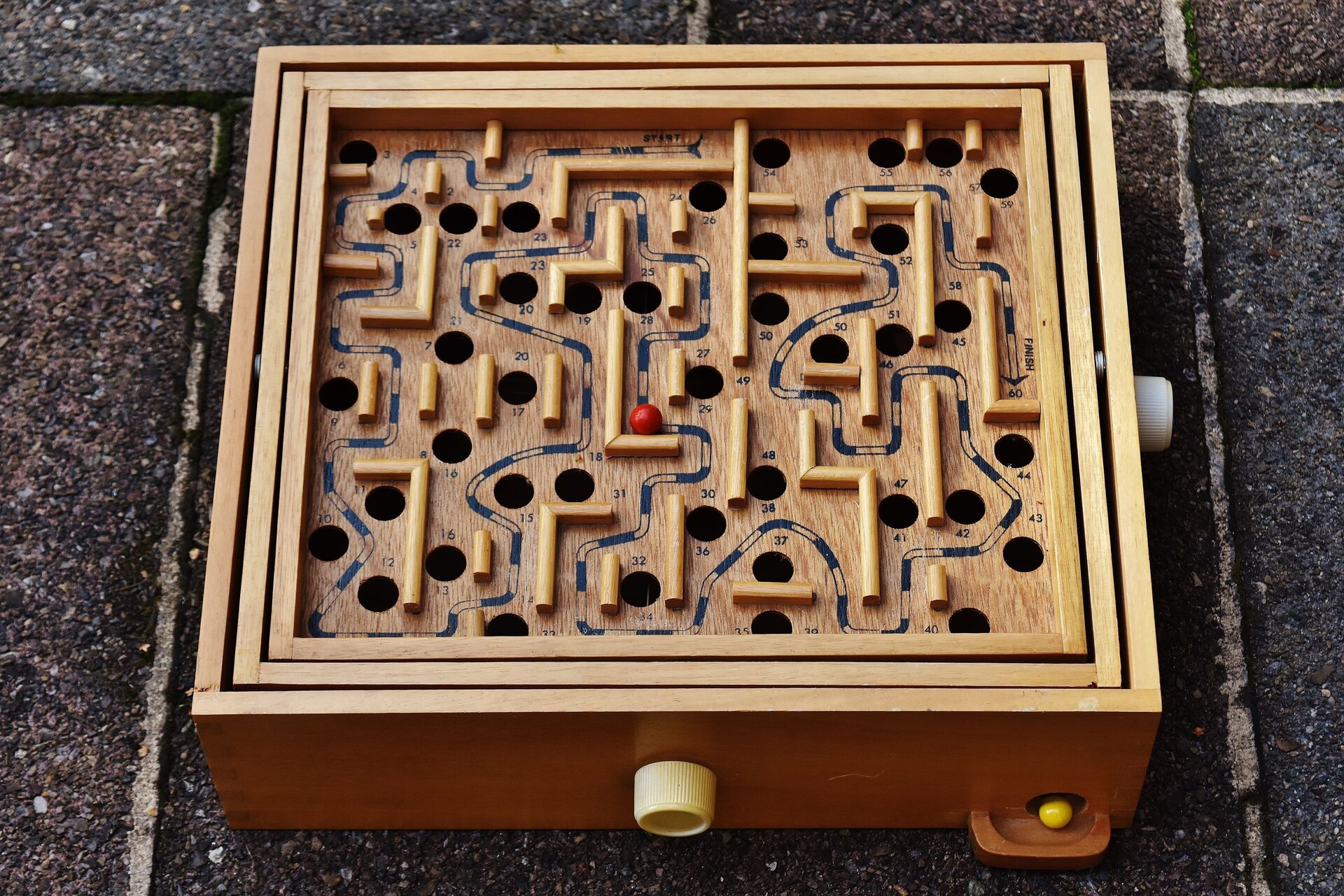 Ilustracja przedstawia drewnianą zabawkę - labirynt z czerwoną kuleczką.