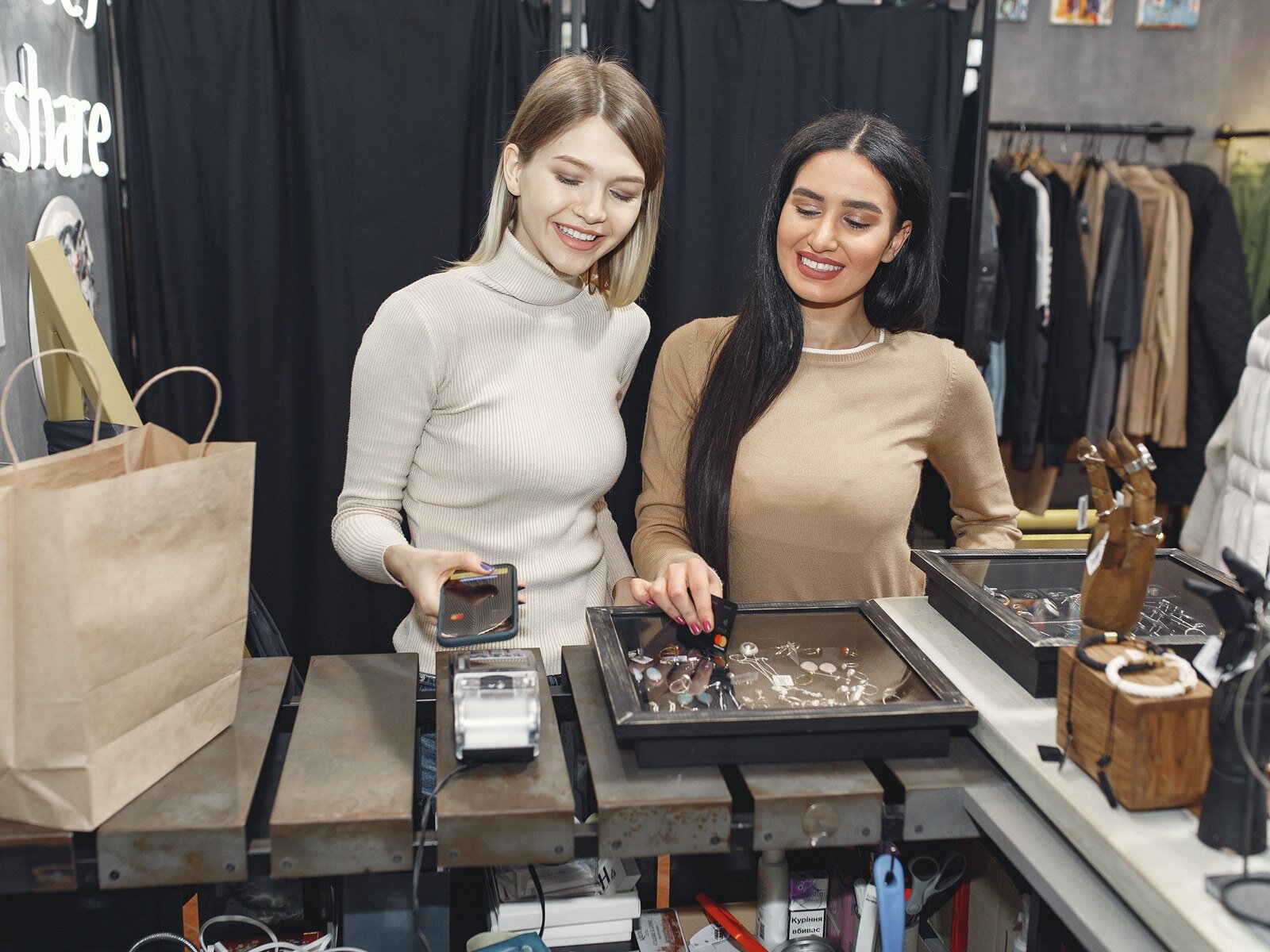 Ilustracja przedstawia dwie młode dziewczyny stojące za ladą w sklepie. Dziewczyna po lewej stronie ma jasne, półdługie włosy, ubrana jest w beżowy golf i uśmiecha się. W prawej ręce trzyma telefon, który zbliża do czytnika kart. Dziewczyna po prawej stronie ma długie czarne włosy, jest ubrana w jasnobrązowy sweter, uśmiecha się, a w prawej dłoni trzyma kartę płatniczą. Na blacie przed nimi widoczne są dwie małe witryny z biżuterią, drewniany stojak w kształcie ręki na pierścionki, duża szara torba zakupowa. W tle widoczne są przymierzalnie przesłonięte czarnymi zasłonami, na prawo od których widoczne są wieszaki z ubraniami.