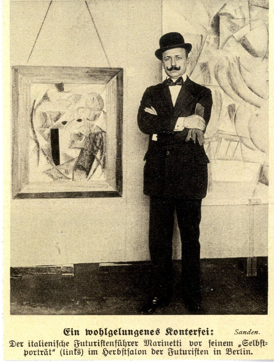 Zdjęcie przedstawia stojącego, dojrzałego mężczyznę. Mężczyzna ma owalną twarz, długie, podkręcone wąsy. Ubrany jest w koszulę, garnitur, ma zawiązaną muchę pod szyją. Na głowie założony melonik, Męzczyzna trzyma skrzyżowane na piersiach ręce, W prawej dłoni ma rękawiczki. Za jego plecami znajduje się ściana, a na niej zawieszone dwa abstrakcyjne obrazy. Pod zdjęciem napis w języku niemieckim.