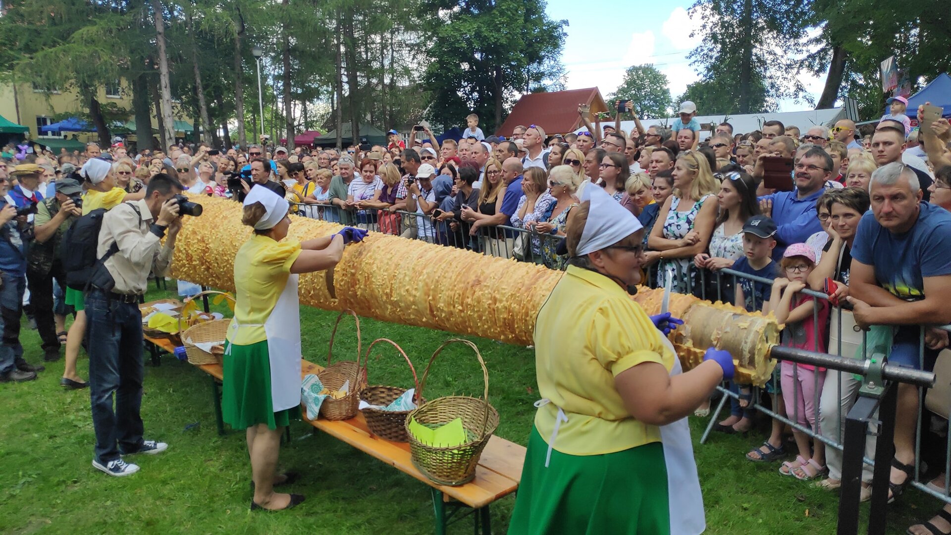Zdjęcie przedstawia kobiety krojące olbrzymiego sękacza i tłum ludzi przyglądający im się. Kobiety te ubrane są w żółte koszulki w krótki rękaw oraz zielone spódniczki do kolan. Mają na sobie ochronny biały fartuch, białe chusty na głowie oraz  niebieskie rękawiczki jednorazowe na dłoniach w których trzymają nóż. Przy nogach stoi ławka na której znajdują się wiklinowe koszyki. Za kobietami stoi mężczyzna , który fotografuje wydarzenie. Tłum ludzi stojący  naprzeciwko za barierkami ubrany jest w lekką wiosenną odzież, próbuje uwiecznić chwile  z wydarzenia przy użyciu telefonów komórkowych. 