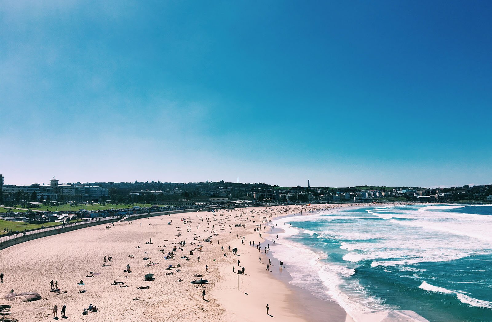 Zdjęcie przedstawia szeroką, piaszczystą plażę, która ciągnie się po łuku skręcającym w prawo. Na plaży widoczni są siedzący i stojący ludzie. Na prawo od plaży widoczny jest zbiornik wodny z zielono‑granatową wodą oraz spienionymi, białymi falami płynącymi w kierunku plaży. Na lewo od plaży widoczny jest pas lądu porośniętego zieloną trawą, z licznymi budynkami usytuowanymi wzdłuż wybrzeża. U góry widoczne jest błękitne niebo.
