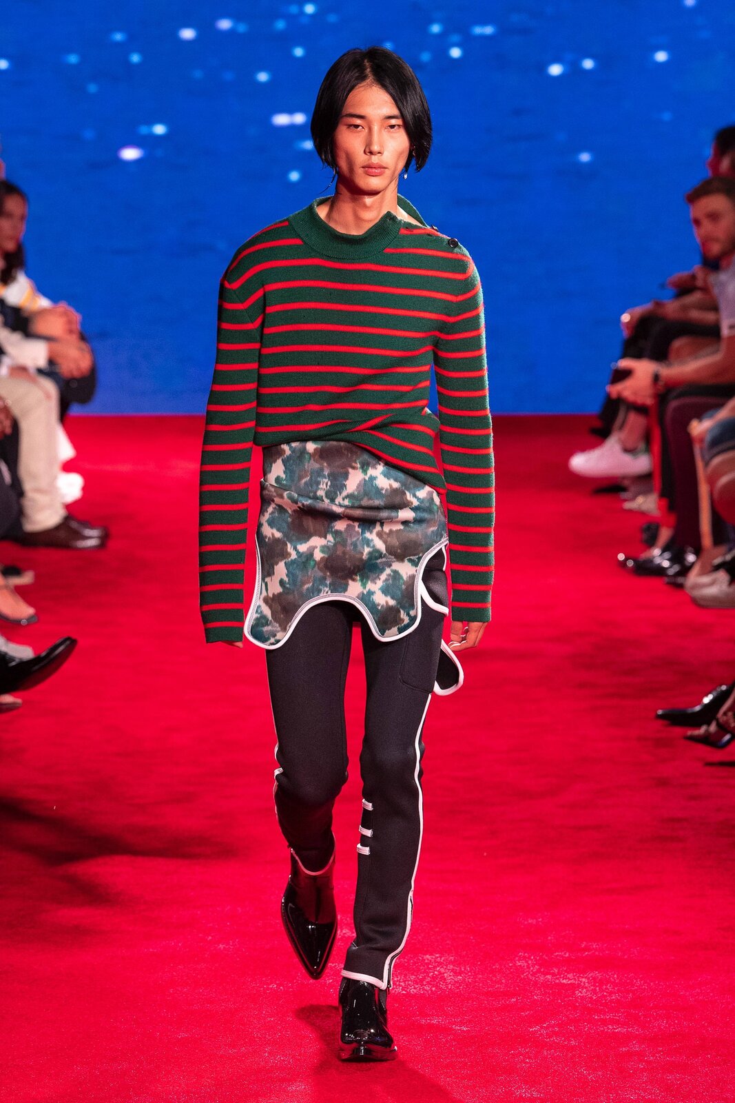 Fotografia przedstawia modela na wybiegu. Ma on na sobie czerwono‑czarną bluzkę w paski, czarne spodnie z białymi liniami po bokach oraz lakierowane buty ze spiczastymi czubkami. Pomiędzy jego bluzką a spodniami widoczny jest element materiału we wzór moro. W tle widać  siedzących ludzi. Dywan, po którym chodzi model, jest czerwony.