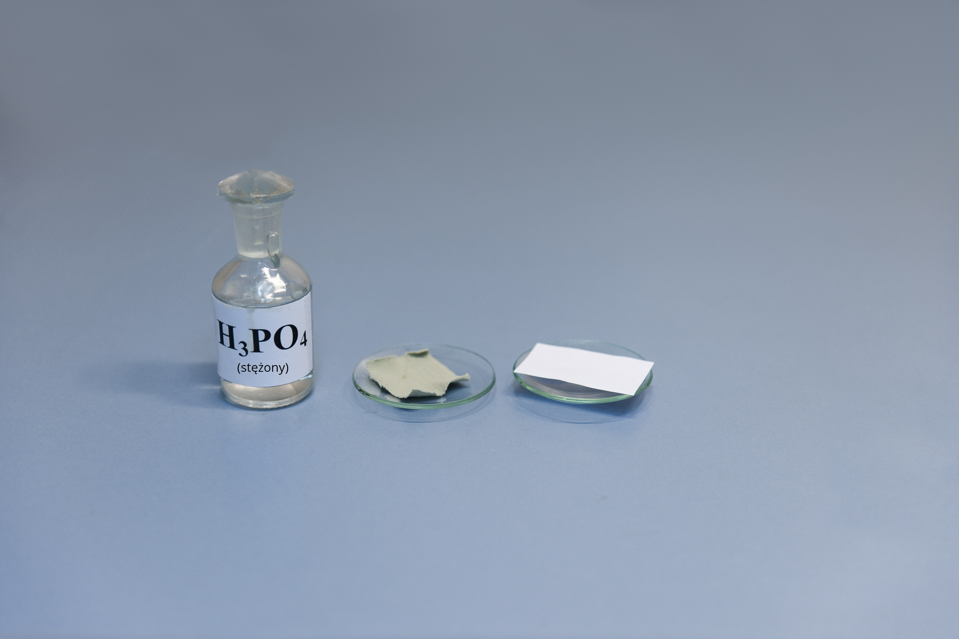 Pierwsze zdjęcie w galerii. Na ilustracji zaprezentowana jest szklana butelka z etykietką i napisem H3PO4 stojąca na niebieskim podłożu. Po jej prawej stronie na blacie laboratoryjnym stoją w szeregu dwa szkiełka zegarkowe, na których położono kolejno kawałek tkaniny i skrawek papieru.