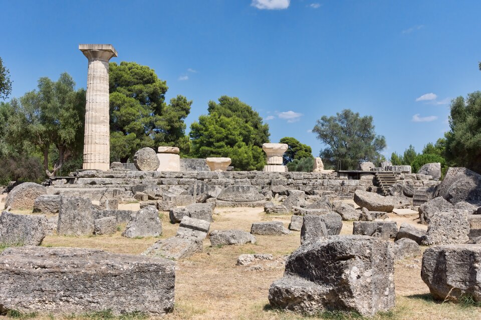 Ilustracja przedstawia ruiny świątyni Zeusa Olimpijskiego na terenie archeologicznym. Na pierwszym planie widoczne są duże, nieregularne bloki kamienne leżące na ziemi. Po lewej stronie znajdują się fragmenty przewróconych kolumn ułożonych poziomo. Po prawej stronie stoi jedna zrekonstruowana kolumna, wysoka i pionowa, z wyraźnie zaznaczonymi żłobieniami. W tle rosną gęste, zielone drzewa, a nad nimi rozciąga się jasne, bezchmurne niebo.