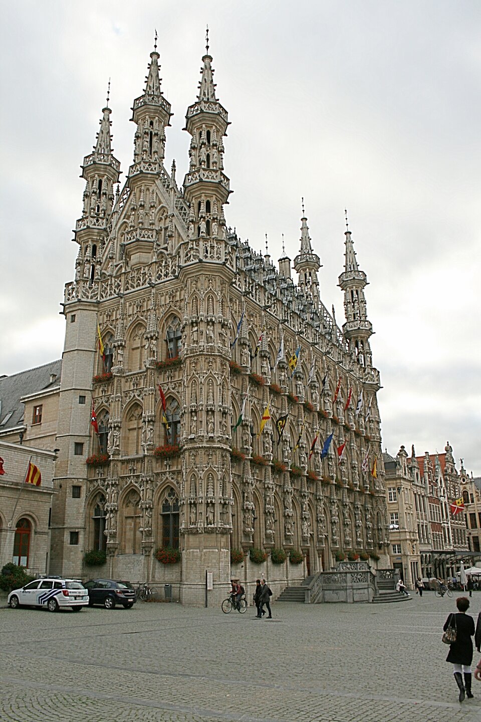 Fotografia przedstawia Ratusz w Leuven. Jest to budowla trzypiętrowa zbudowana z jasnego kamienia, zwieńczona balustradą, przykryta dachem dwuspadowym z licznymi lukarnami. Z każdego ze szczytów wyrastają po trzy smukłe wieżyczki, zwieńczone stożkowymi hełmami. Fasady, szczyty i wieżyczki są niezwykle bogato zdobione za pomocą laskowań i nisz, w których umieszczono posągi sławnych postaci, zasłużonych dla miasta Leuven. W najniższym rzędzie, na parterze, umieszczono figury uczonych, artystów i możnych mieszkańców Leuven. Pierwsze piętro obsadzono figurami symbolizującymi wolności miejskie i świętych patronów lokalnych parafii. Drugie piętro prezentuje postaci wszystkich władców miasta – hrabiów Leuven i książąt Brabancji. Na trzecim piętrze znajdują się rzeźby hrabiów i książąt Leuven z różnych wieków. Główna fasada ma schody wejściowe, prowadzące do dwóch portali, powyżej których umieszczono figury św. Piotra (z lewej) i Madonny z Dzieciątkiem (z prawej). Budynek jest smukły i strzelisty. 