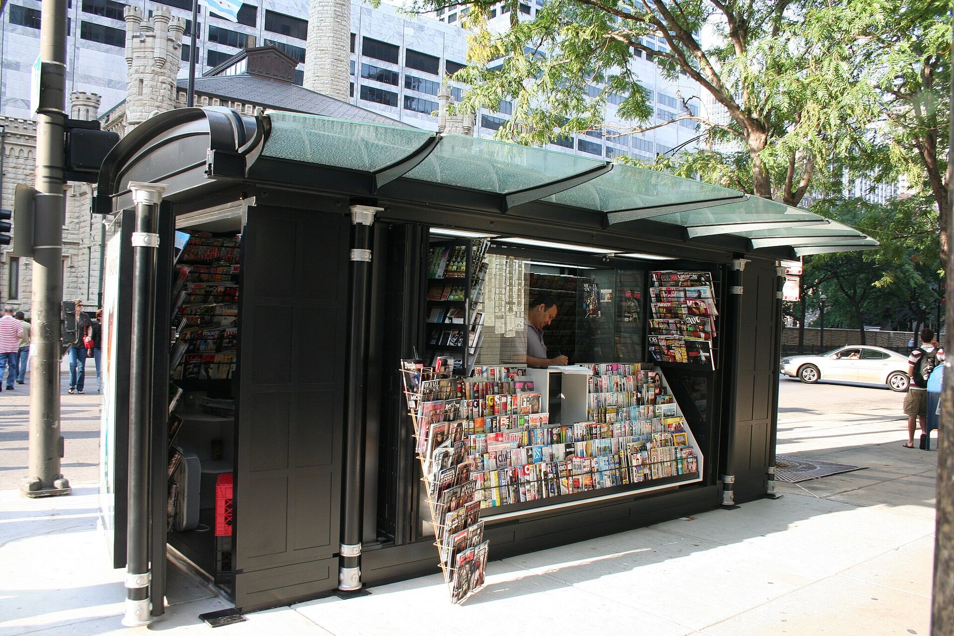 Zdjęcie przedstawia stoisko z gazetami, gdzie możemy również znaleźć pocztówki i płyty. Cała przednia ściana jest obwieszona różnymi gazetami i czasopismami. Przed nimi jest stojak z pocztówkami, a wewnątrz stoiska stoi mężczyzna.