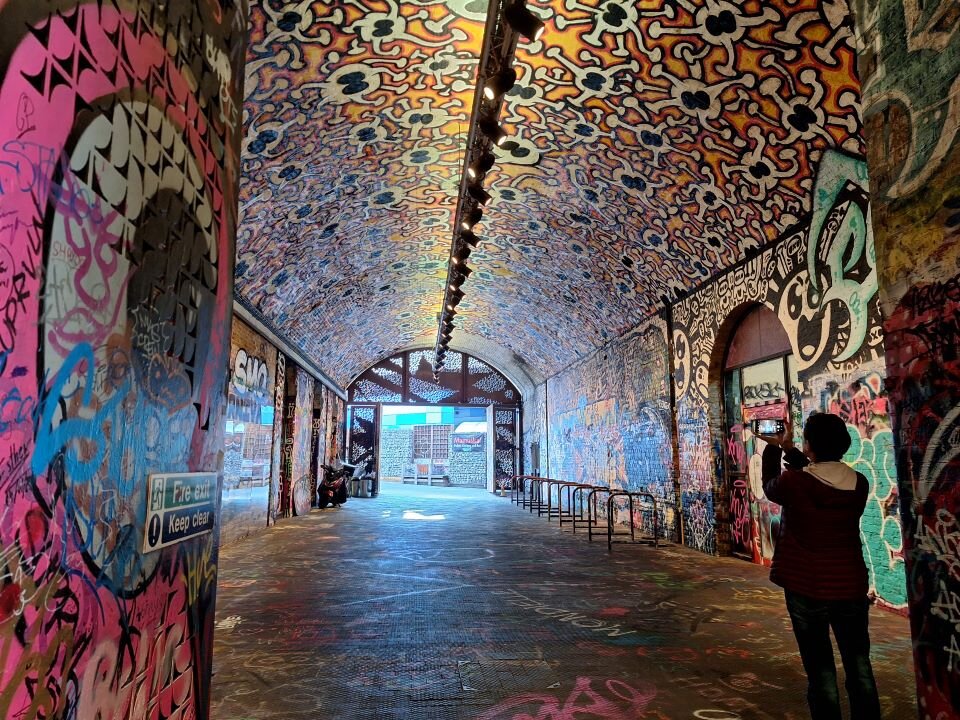 Ilustracja przedstawia fotografię wnętrza tunelu w perspektywie. Wszystkie ściany tunelu wraz z sufitem jest pokryty wielobarwnymi malowidłami. Z prawej strony na pierwszym planie jest widoczna stojąca tyłem postać, która wykonuje zdjęcie tunelu. W oddali widać wyjście z tunelu. Fotografia bardzo dobrze obrazuje perspektywę zbieżną.