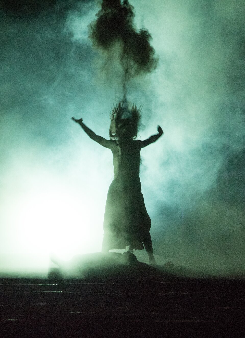 Zdjęcie przedstawia scenę teatralną. Stoi na niej spowita dymem postać kobiety. U jej stóp leży człowiek.