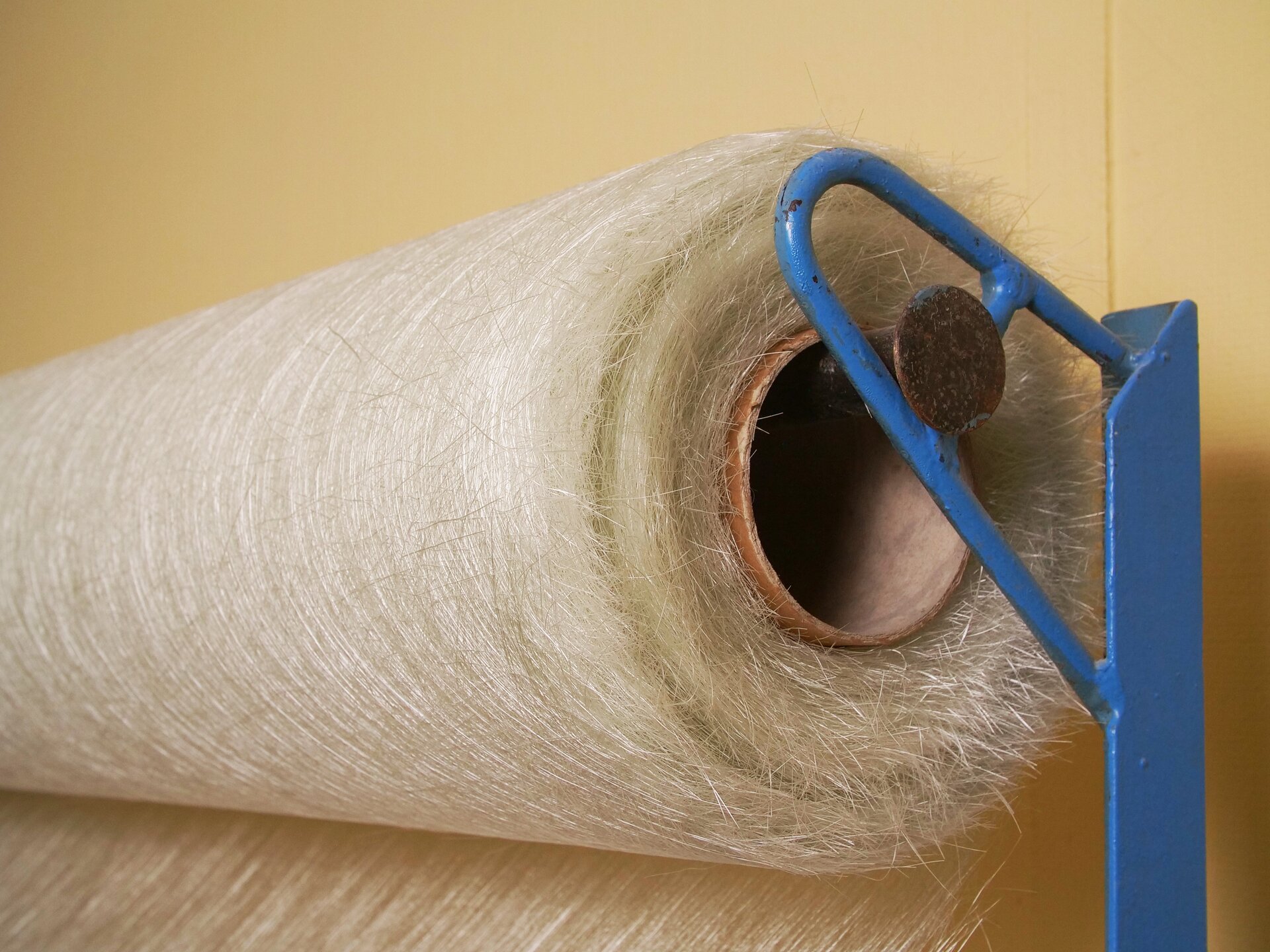 Zdjęcie przedstawia watę szklaną. Jest ona nawinięta na szpulę zawieszoną poziomo na niebieskim metalowym elemencie. Wata ma postać giętkiego arkusza o jasnym, białawym zabarwieniu. Jej struktura zbudowana jest ze sprasowanych włókien szklanych.