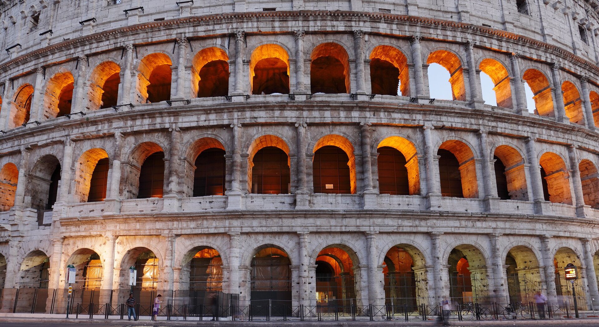 Ilustracja przedstawia rzymskie Koloseum - amfiteatr w Rzymie we Włoszech. Na fotografii widzimy budowlę, która nie mieści się w kadrze. Na każdym z trzech pięter zaokrąglonej bryły amfiteatru znajduje się ciąg rytmicznie rozmieszczonych arkad. Na najniższej kondygnacji zastosowano porządek toskański, na drugiej joński, na trzeciej – koryncki. Wnętrze budowli jest oświetlone. 