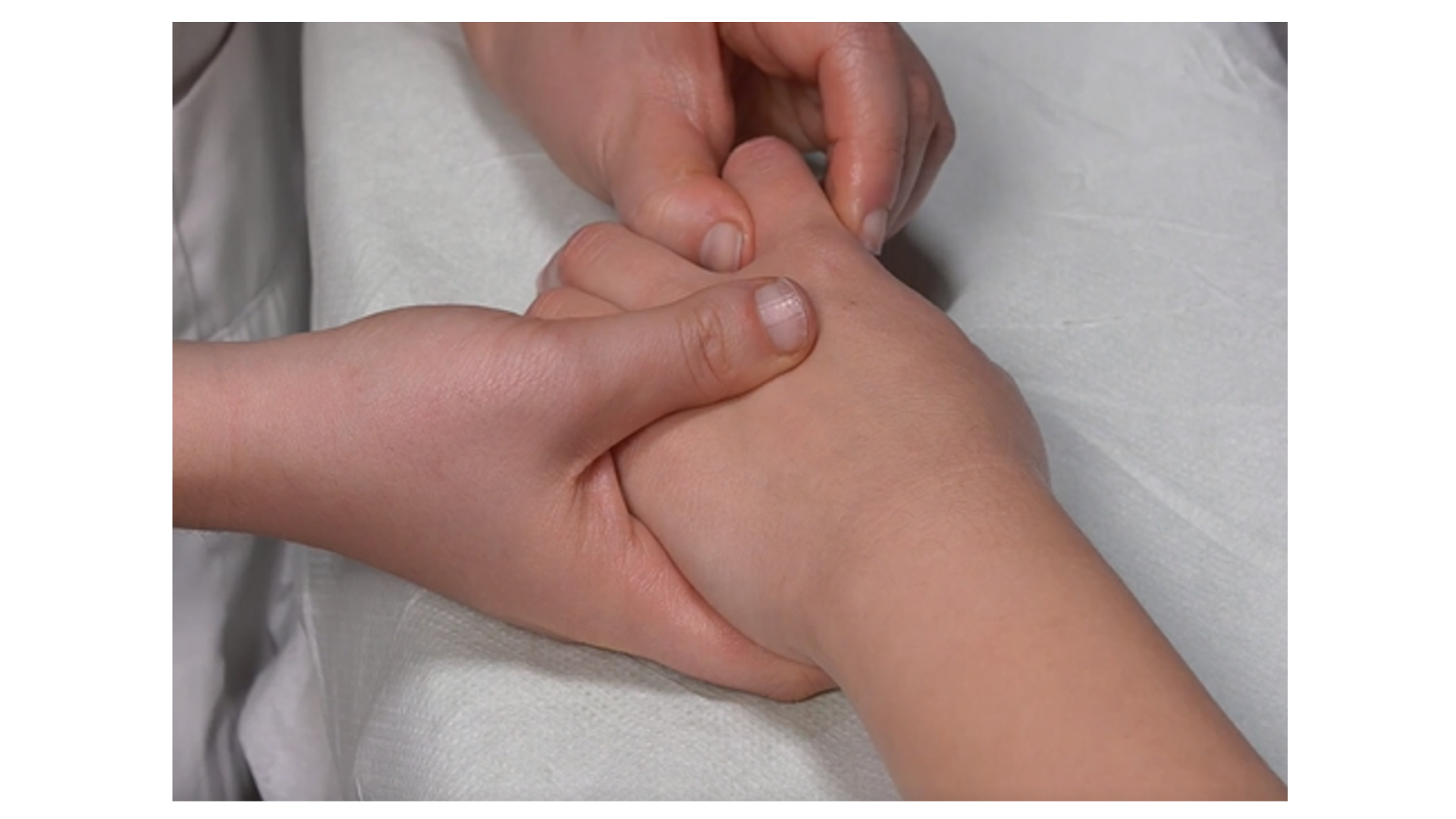 Zdjęcie przedstawia lewą rękę pacjenta leżącą na stole do masażu. Masażysta uciska kciukami kości palca wskazującego pacjenta. Drugą ręką przytrzymuje rękę pacjenta. 