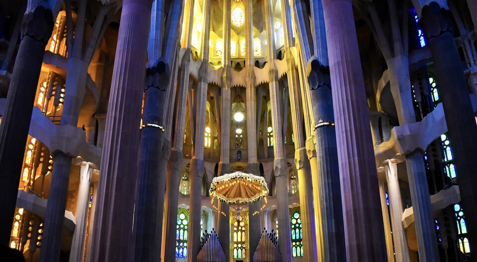 Ilustracja przedstawia Wnętrze katedry Sagrada Família. Ukazuje centralną część prezbiterium z wysokimi, rozgałęziającymi się kolumnami, które unoszą sklepienie o złożonych, geometrycznych formach. Witraże w odcieniach zieleni, żółci i błękitu przepuszczają światło, tworząc na kamiennych powierzchniach miękkie, kolorowe refleksy. W centrum widoczny jest geometryczny żyrandol z wizerunkiem Chrystusa u dołu.