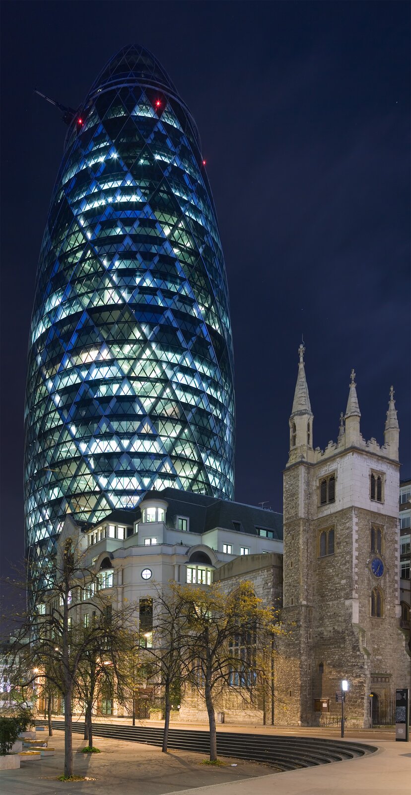 Fotografia przedstawia biurowiec w Londynie „The Gherkin”. Ma kształt walca rozszerzającego się pośrodku. Jego zewnętrzna konstrukcja zbudowana jest z krzyżujących się ze sobą pod ukosem linii, które tworzą romby. Okna budynku są oświetlone. Na pierwszym planie widoczne są zabudowania z wieżą zbudowane z jasnego kamienia oraz kamienica z białą fasadą. 