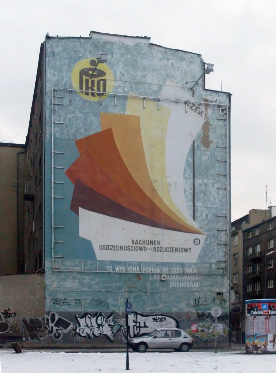 Zdjęcie przedstawia mural, stanowiący dawną reklamę banku PKO. Na budynku widnieje napis „Rachunek oszczędnościowo‑rozliczeniowy”, nad którym ułożone są druki. W górnej części, po lewej stronie znajduje się logo banku: napis PKO na tle żółtego koła. W dolnej części budynku zamieszczone są inne, czarno‑białe graffiti.