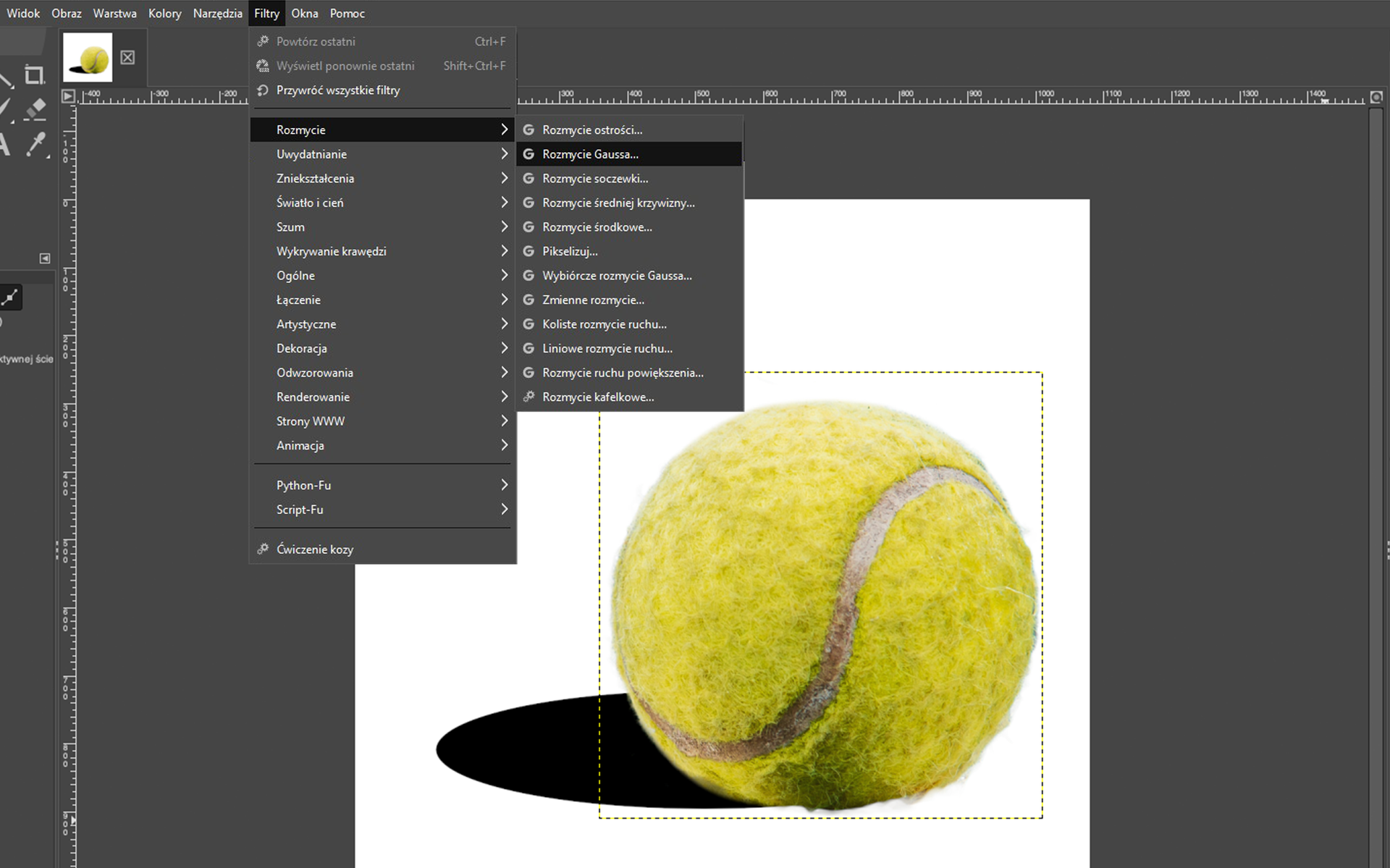 Ilustracja przedstawia okno programu. Z menu wybrano zakładkę Filtry, tam z listy wybrano: Rozmycie, a kolejno: Rozmycie Gaussa. W obszarze roboczym jest piłka tenisowa na białym tle. W dolnej części piłki po lewej stronie jest część czarnej elipsy. Przypomina cień.  