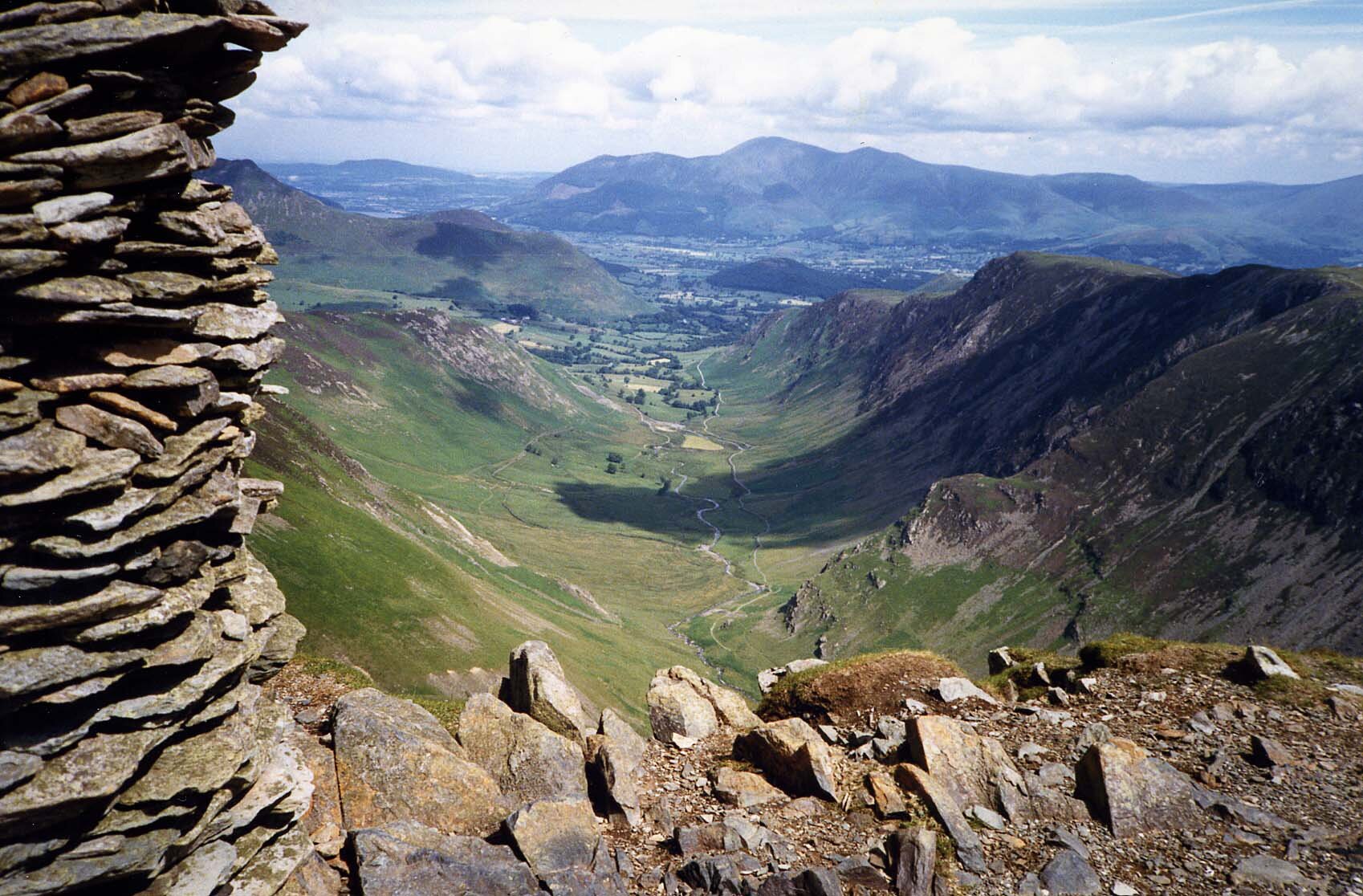 Fotografia przedstawia widok na dolinę znajdującą się pomiędzy wzgórzami. Zdjęcie zostało wykonane z wysokiego punktu, skąd dolina jest dobrze widoczna. W tle inne góry.