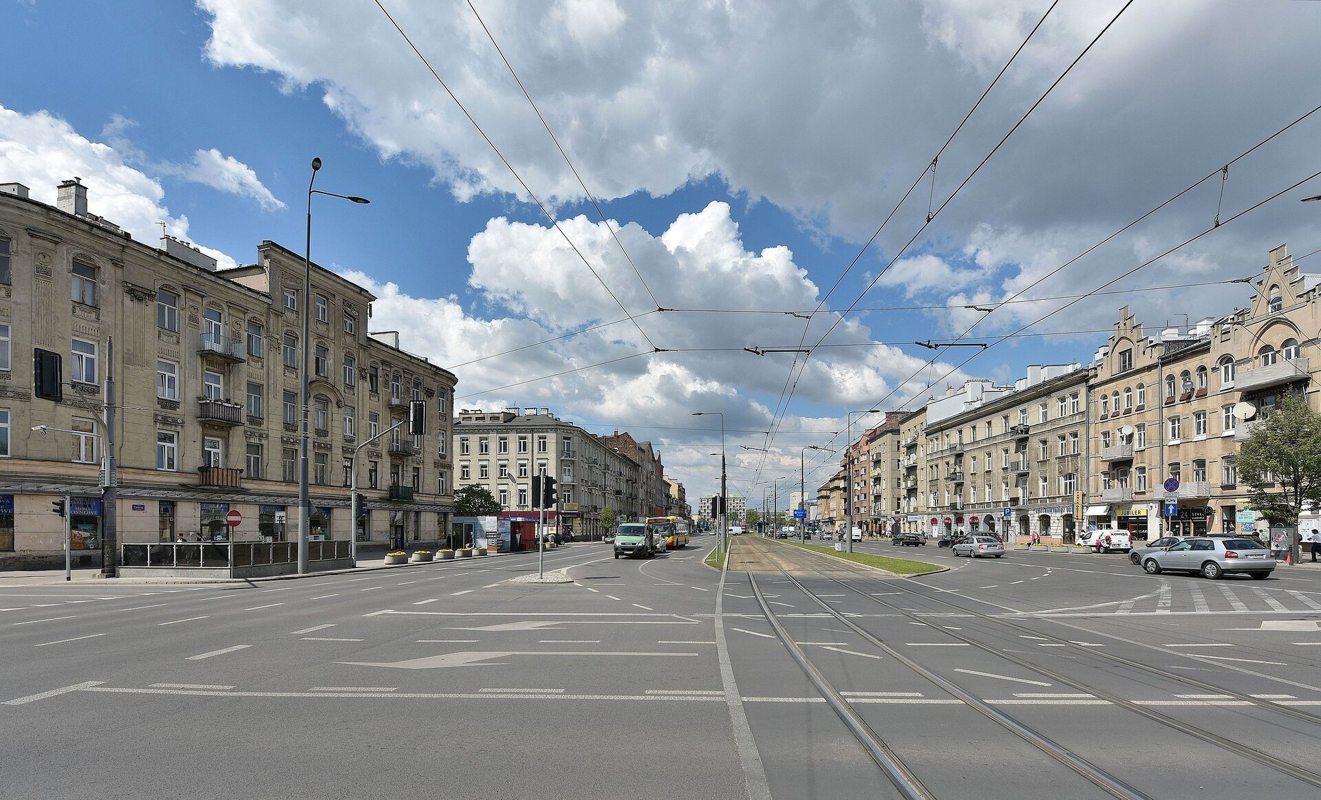 Zdjęcie przedstawia szeroką, kilkupasmową ulicę. Trzy pasy w jednym kierunku i trzy pasy w drugim. Między nimi znajduje się torowisko tramwajowe. Wzdłuż drogi stoją niewysokie kamienice.