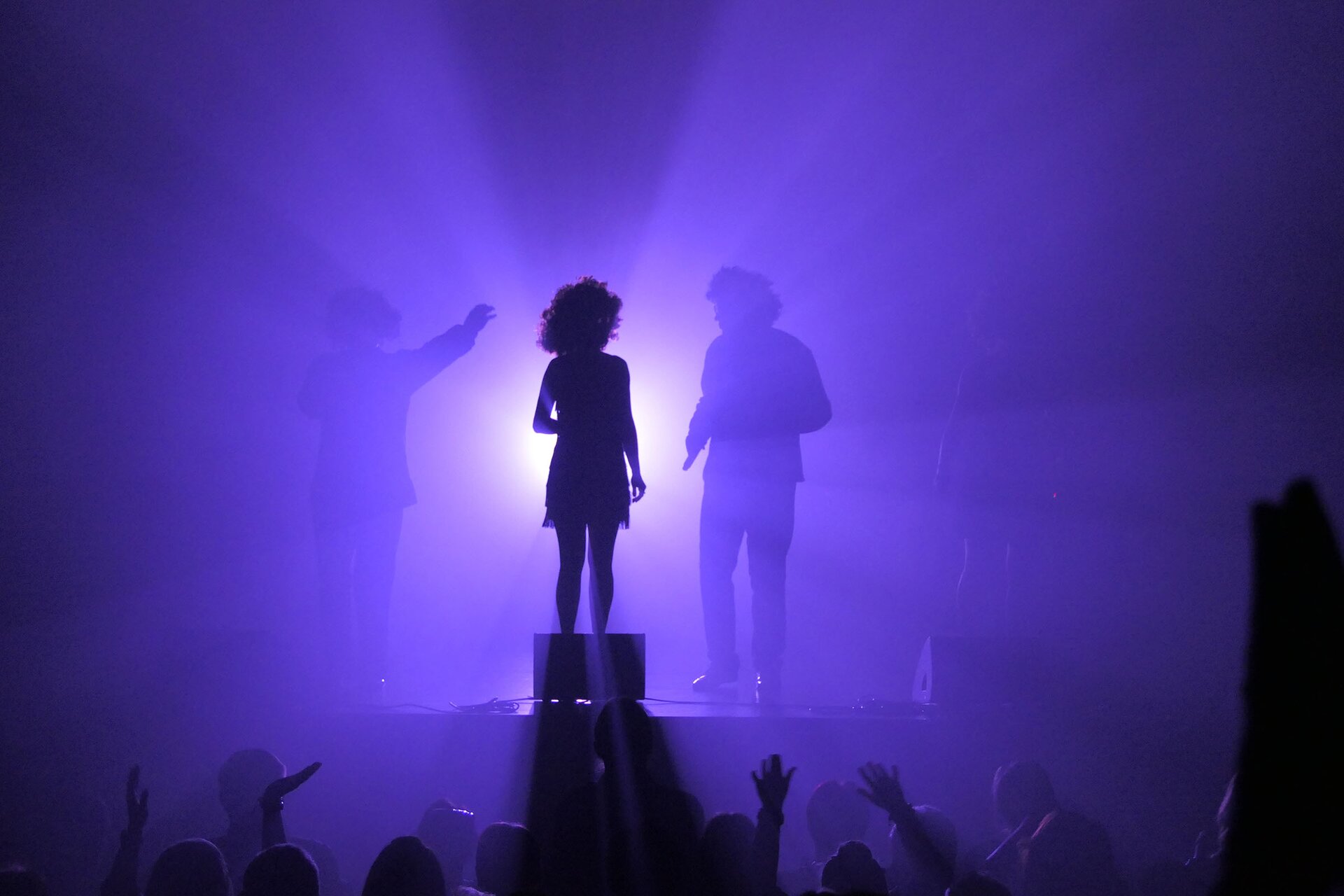 Zdjęcie przedstawia scenę oświetloną na filetowo, na scenie stoi kobieta w sukience, po obu jej stronach stoją mężczyźni. Pod sceną stoją ludzie z wyciągniętymi rękami ku górze.