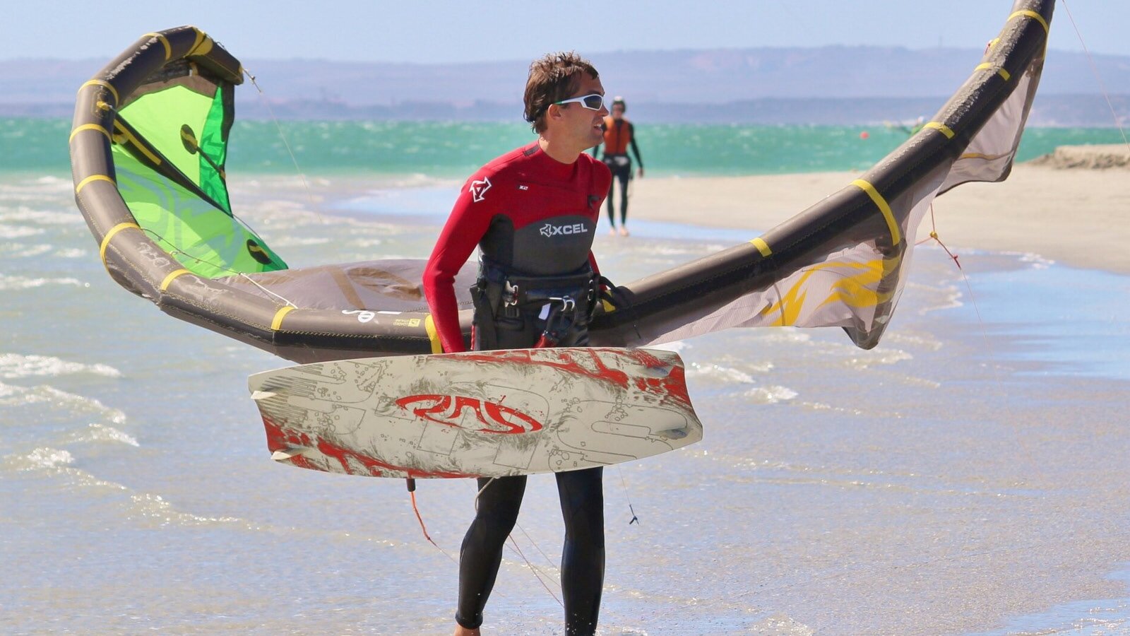 Ilustracja przedstawia scenę, gdy młody mężczyznę w kombinezonie i okularach, który wychodzi z wody na plażę. W jednej ręce trzyma deskę do surfowania, a w drugiej żagiel.
