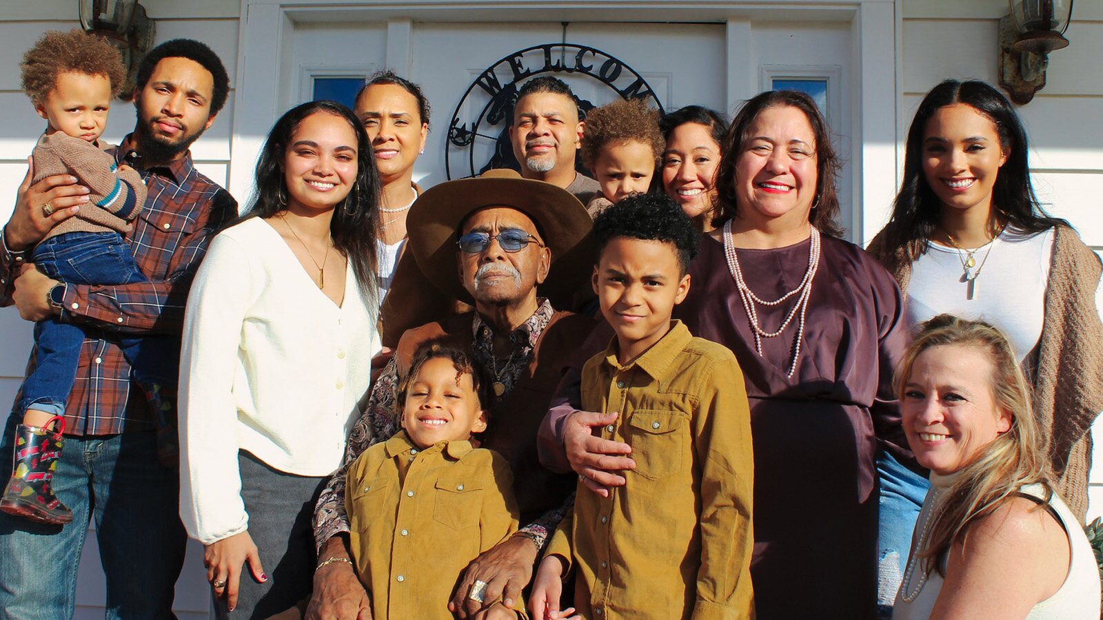 Zdjęcie przedstawia grupę osób w różnym wieku, stojących blisko siebie i pozujących do zdjęcia. Wszyscy patrzą prosto w obiektyw, obejmują się i uśmiechają. W tle widoczne są białe drzwi domu.