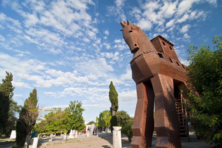 Zdjęcie przedstawia drewnianego konia stojącego na placu, dookoła którego znajdują się niewysokie drzewa i krzewy. Z prawej strony stoi koń zrobiony z drewna, w jego tułowiu znajdują się kwadratowe okna. Na grzbiecie konia znajduje drewniana budka z dwoma kwadratowymi oknami. Między nogami są drewniane schody z barierką prowadzące do brzucha konia. W tle krzewy i niebieskie, usiane delikatnie chmurami niebo.