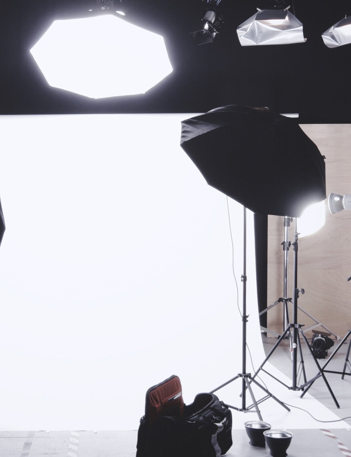 Zdjęcie przedstawia dyfuzor. Widoczne jest pomieszczenie, w którym po prawej stronie widoczne są dwa statywy na których na jednym zamocowany jest czarny parasol, zaś na drugim lampa. U góry widoczny jest mocno oświetlony parasol - dyfuzor światła. W centralnej części mocno oświetlony prostokąt - tło zdjęciowe. Na pierwszym planie zarys czarnej torby fotograficznej.