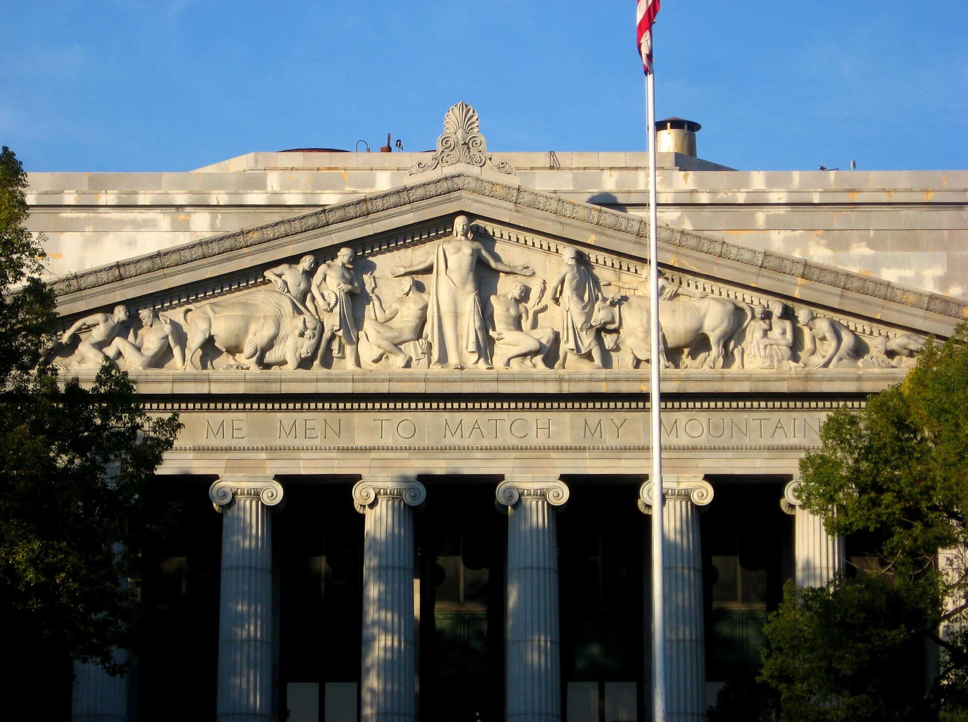 Zdjęcie przedstawia trójkątny tympanon z rzeźbami, który podtrzymywany jest przez okrągłe, żebrowane kolumny. W centralnej części tympanonu stoi półnaga postać z rękoma rozłożonymi na boki. Obok postaci, symetrycznie po obu stronach postacie siedzące. Następnie, po obu stronach znajdują się zwierzęta oraz kolejne postaci ludzi.