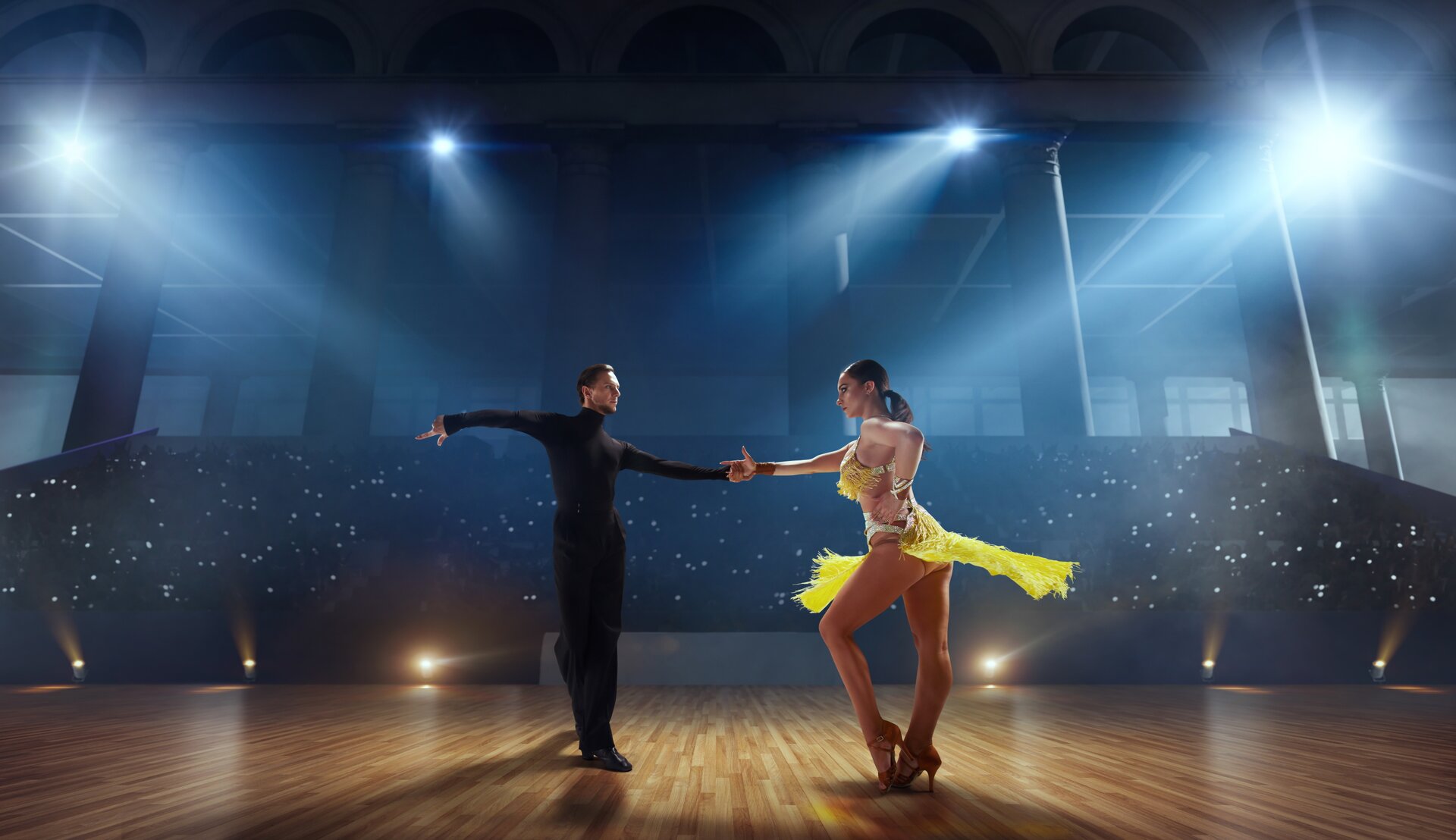 Fotografia przedstawia parę tańczącą Cha‑chę – latynoamerykański taniec, na konkursie tańca. Na pierwszym planie widoczne są dwie osoby – mężczyzna i kobieta. Mężczyzna ubrany jest w ciemną bluzkę z długim rękawem oraz golfem, a także czarne spodnie. Ma krótkie, zaczesane do tyłu włosy. Jedną rękę skierował do tyłu na wysokość barków, drugą trzyma prawą dłoń stojącej na przeciwko niego kobiety, której długie, ciemne włosy spięte są w niski kucyk. Jej lewa ręka spoczywa na jej odsłoniętej talii. Nogi tancerki są ustawione w rozkroku. Lewa noga jest zgięta w kolanie, a prawa jest wyprostowana. Kobieta ma na sobie żółty komplet – top oraz spódnicę, ze srebrnymi pasami, które utrzymują strój na ciele kobiety. W tle widoczne są trybuny oraz lampy rozświetlające drewniany parkiet. 