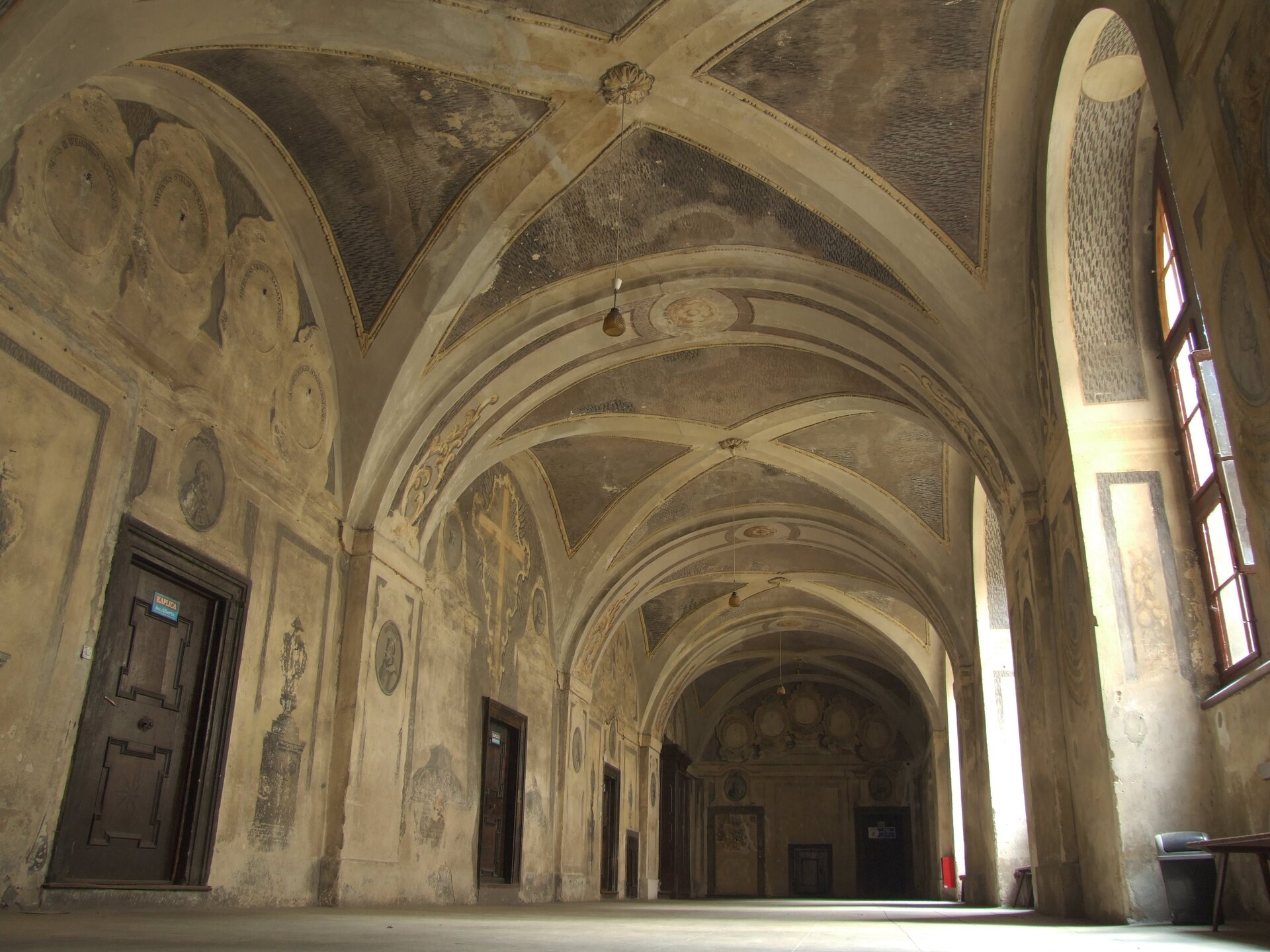 Zdjęcie przedstawia szeroki korytarz, po lewej stronie znajduje się kilka par drzwi, a po prawej duże, okna. Sufit ma charakterystyczne, łukowate sklepienie. Na ścianach widnieją wyblakłe, sakralne malowidła.
